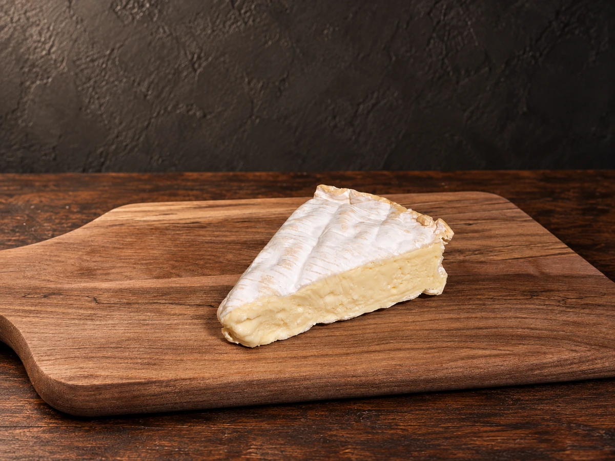 Een ronde Camembert Rustique met een witte, fluweelachtige schimmelkorst, liggend op een houten plank met enkele kersen en walnoten ernaast.